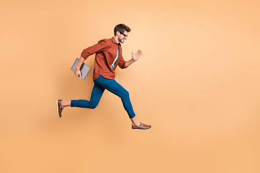 Full length body size view of his he nice attractive cheerful cheery successful brunet, guy jumping in air carrying laptop running fast late hurry-up isolated over beige color pastel background