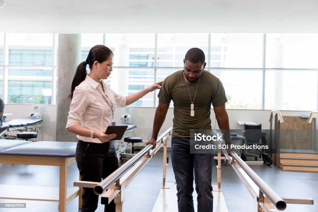 Physical therapist guides military vet as he uses injured foot The female physical therapist guides the mid adult male military veteran as he walks on his foot during therapy. Veteran Stock Photo
