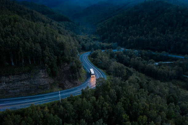 vista aérea de un giro brusco en un camino de montaña entre árboles verdes del bosque. semicamión con remolque de carga y faros brillantes en una carretera oscura - driving night car headlight fotografías e imágenes de stock