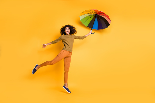 Full length body size photo of wavy cheerful cute nice charming pretty girlfriend, flying with umbrella wearing orange pants trousers footwear isolated over vivid yellow color background