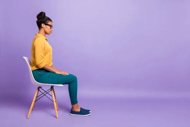 Photo of Full length profile photo of pretty dark skin lady on chair look empty space listen employer question interview wear specs yellow shirt trousers isolated purple color background