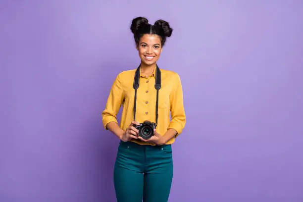 Photo of Photo of amazing dark skin lady holding photo digicam in hands positive cheerful mood photographer wear yellow shirt trousers isolated purple color background