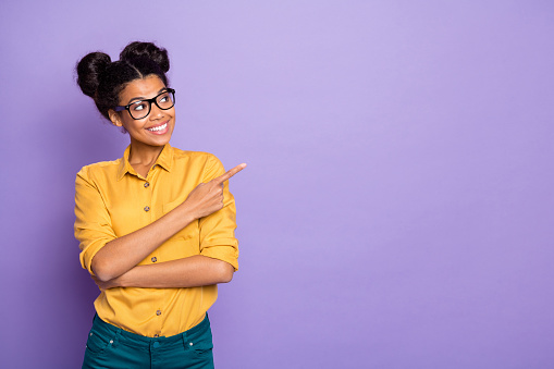 Photo of amazing dark skin lady indicating finger to empty space advising novelty interested looking side wear specs yellow shirt isolated purple color background