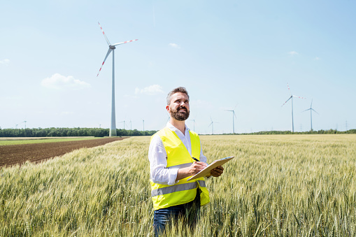 An engineer or technician with clipboard standing on a field on wind farm, making notes.