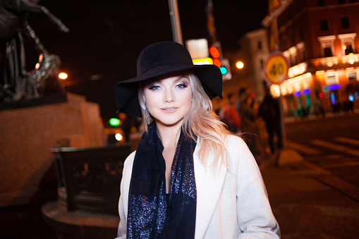 Stylish and elegant young woman in a beige coat and black hat on the city . Night city in autumn. Portrait of charming blonde at night outdoors, city lights at night.