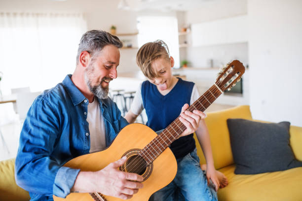 Mature father with small son sitting on sofa indoors, playing guitar. A mature father with small son sitting on sofa indoors, playing guitar.. father and son guitar stock pictures, royalty-free photos & images