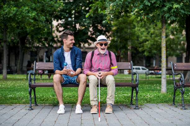 Young man and blind senior with white cane sitting on bench in park in city. A young man and blind senior with white cane sitting on bench in park in city. blind persons cane stock pictures, royalty-free photos & images