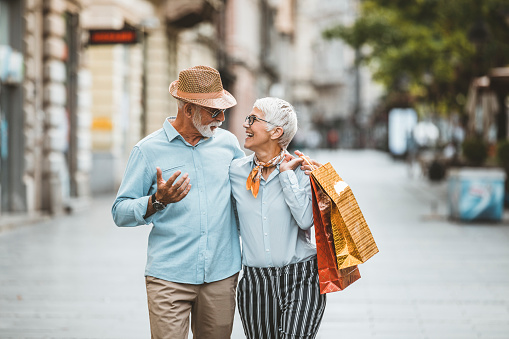 Senior Couple, Casually Dressed, is Walking in the City Center with Shopping Bags. Mature Handsome Man with Hat and Sunglasses and Grey Beard is Walking with his Beautiful Wife with Grey Hair and Talking About the Successful Shopping.