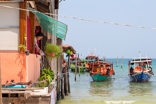 Koh Rong island, Cambodia - 07 April 2018: sea view with boat, house on piles and woman watering flowers. Everyday life of tropical island. Cambodian traditional boat and house. Cambodian lifestyle