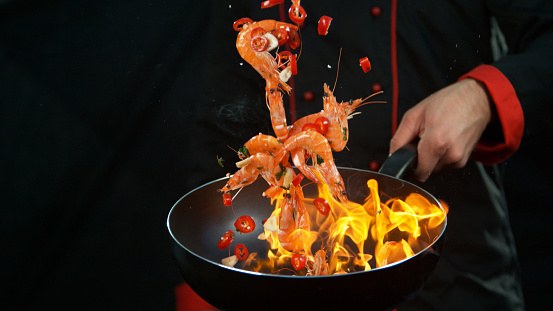 Closeup of chef holding wok pan with falling prawns with chilli peppers. Isolated on black background