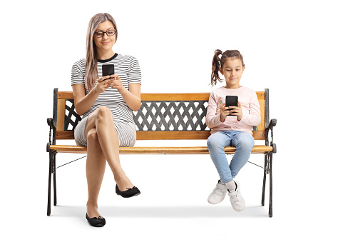 Full length shot of a mother and child sitting on a bench and using mobile phones isolated on white background