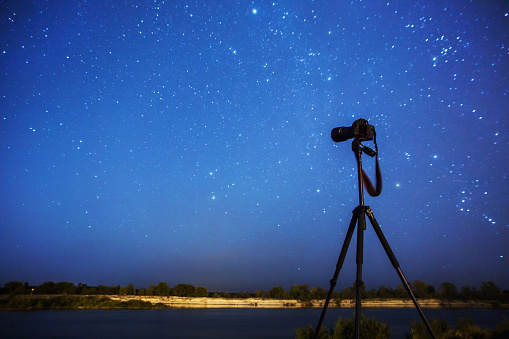 Night photography. Silhouette of camera on tripod on background of starry sky. Long exposure. Many stars in night blue sky. Dark starry landscape. Shooting equipment. Take picture. Professional camera