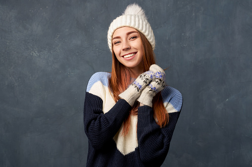 Horizontal shot of a cheerful woman holding her hands to her face. Young girl with ginger long hair wearing warm blue sweater knit cap and woolen mittens isolated on textured wall in Studio.