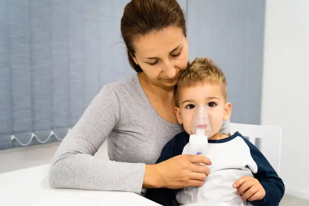 Photo of Woman mother hold her small son boy child using vapor steam inhaler nebulizer mask inhalation at home or hospital medical procedure medicament treatment asthma pneumonia bronchitis coughing sick