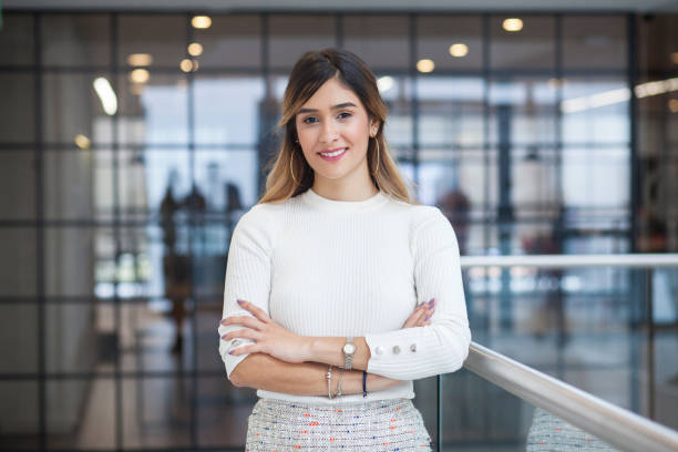 Latin-haired Latina woman with her arms crossed looking at the camera while smiling Executive Latin woman with long brown hair and white skirt and gray skirt with an approximate age of 28 is crossed arms and smiling at the camera with a half-sided stance in one of the corridors of the company for which she works next her a silver railing colombian ethnicity stock pictures, royalty-free photos & images