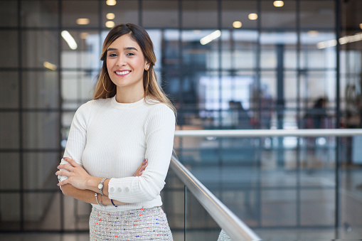 Executive Latin woman with long brown hair and white skirt and gray skirt with an approximate age of 28 is crossed arms and smiling at the camera with a half-sided stance in one of the corridors of the company for which she works next her a silver railing