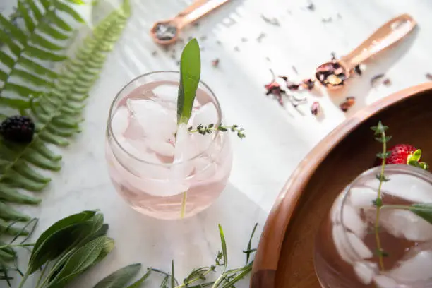Pink artisan cocktail on marble background with botanical herbs and greenery