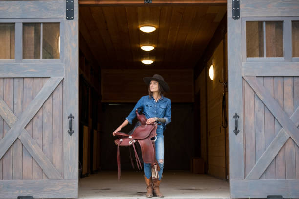 Female rancher holding her saddle in the tacking room. She is walking out the barn towards her horse