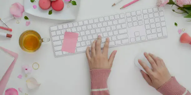 Photo of Overhead shot of young girl typing on keyboard in sweet pink feminine workspace with make up