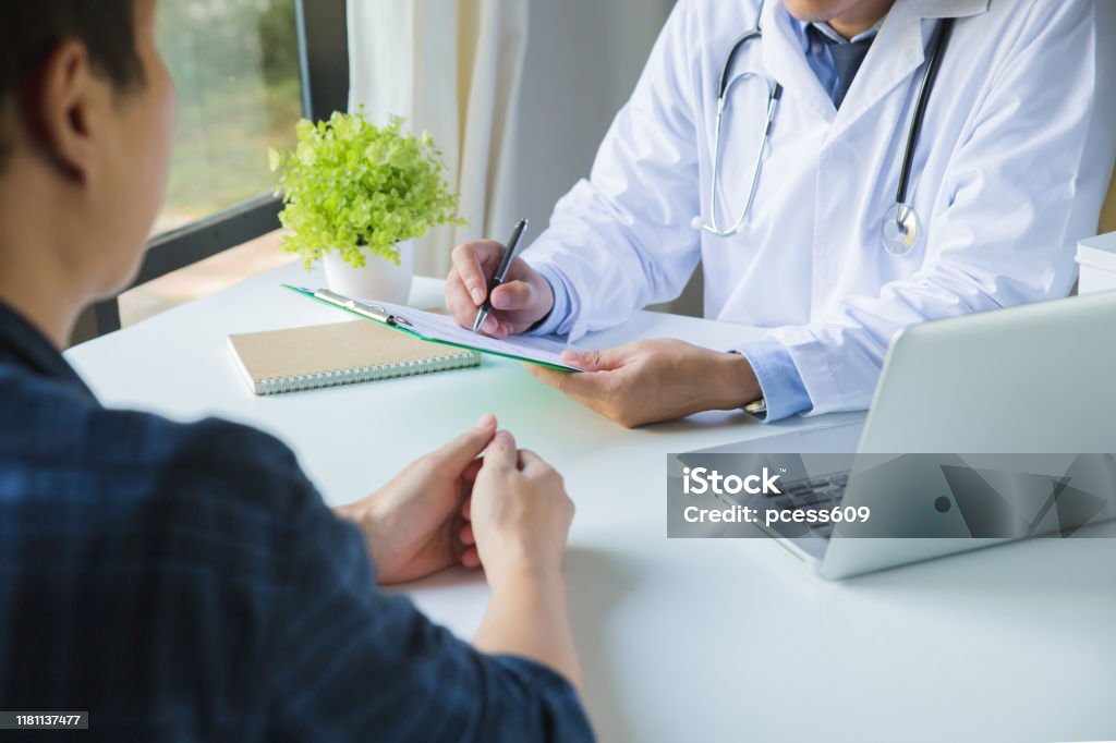 Doctor using a clipboard to fill out a medical history of a young man's medication and patient discussing the results of a physical examination in a clinic Patient Stock Photo