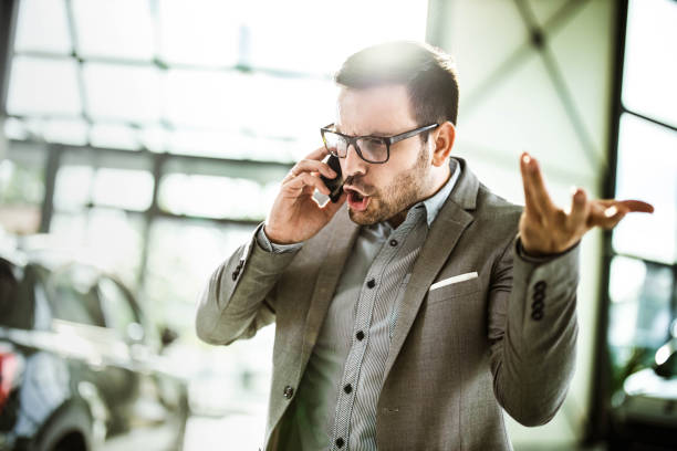 Angry businessman talking on cell phone in a car showroom. Young displeased businessman arguing with someone over smart phone while being in a car showroom. displeased stock pictures, royalty-free photos & images