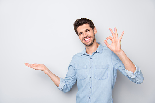 Portrait of positive cheerful promoter man hold hand demonstrate, adverts present feedback recommend perfect novelty show ok sign wear casual style clothes isolated over grey color background