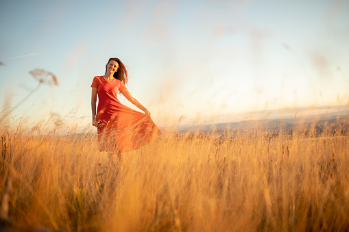 Beautiful mature woman wearing long red dress and enjoying in high grass on mountain at sunset light. She is very happy and cheerful