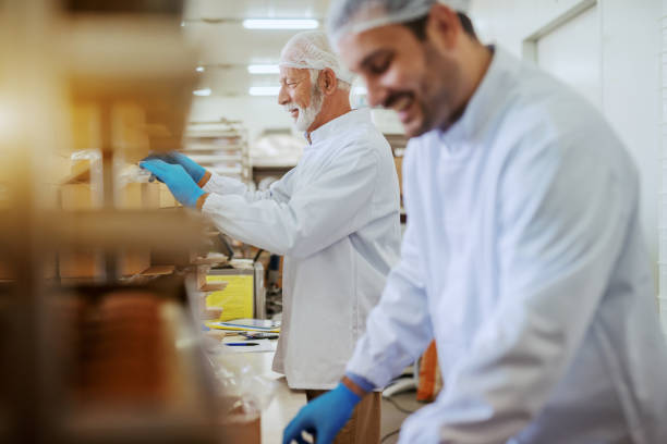 istock Cheerful Caucasian employees dressed in white sterile uniforms packing cookies in boxes while standing in food plant. 1181188198