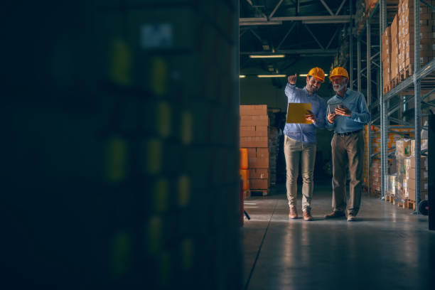 istock CEO going around warehouse with supervisor and talking analyzing sale statistics. Younger man holding folder with data and pointing at boxes while older one holding tablet. Both having yellow helmets. 1181190297