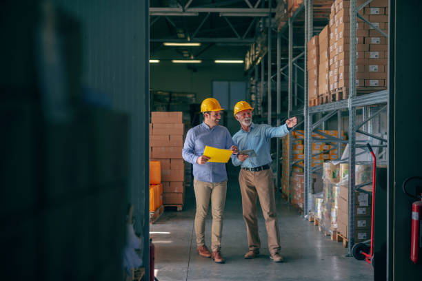 istock CEO going around warehouse with supervisor and talking analyzing sale statistics. Younger man holding folder with data while older one holding tablet and pointing at boxes. Both having yellow helmets. 1181190567