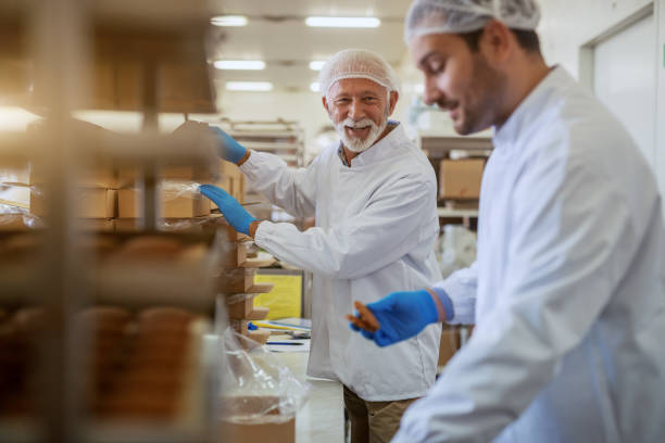 istock Cheerful Caucasian employees dressed in white sterile uniforms packing cookies in boxes while standing in food plant. 1181192005