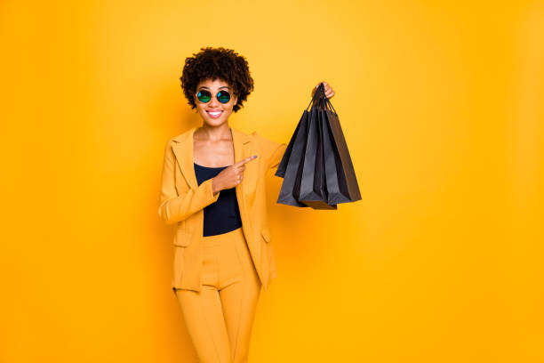 schauen sie echte schnäppchen. porträt der positiven brünette lockige haare afro amerikanische mädchen halten schwarze taschen punkt zeigefinger zeigen, was sie gekauft gekauft tragen stil hose outfit isoliert gelb farbe hintergrund - store suit commercial sign marketing stock-fotos und bilder