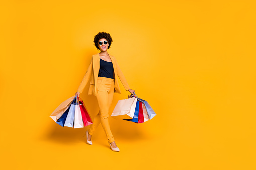 Full body foto de positiva alegre chica afro americana en viaje viaje ir compras encontrado descuentos de venta mantener bolsos usan moda estilo traje pantalones de gafas de sol de tacón aislado fondo de color amarillo aislado photo