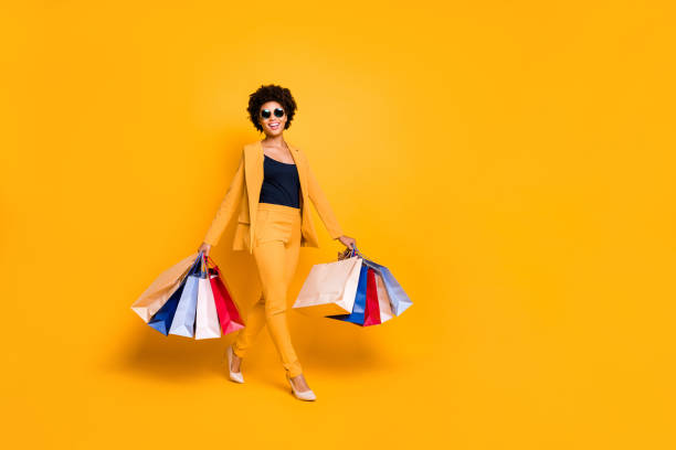 full body photo of positive cheerful afro american girl on travel trip go shopping found sales discounts hold bags wear vogue style outfit sunglass pants high-heels isolated yellow color background - fato de senhora imagens e fotografias de stock