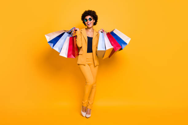 full length photo of positive bruntte hair dark skin girl feel enthusiastic hold bags addicted shopper shopping on leisure time wear style outfit sunglass high-heels isolated yellow color background - shopping bag imagens e fotografias de stock