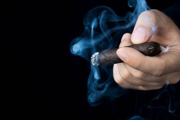 A hand holds a smoldering cigar in smoke against a dark background. Left space for text. Photo of a hand holds a smoldering cigar in smoke against a dark background. Left space for text. man cigar stock pictures, royalty-free photos & images