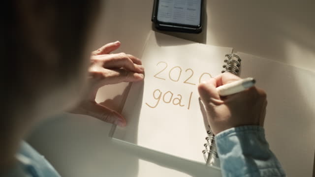Close up woman hand writing 2020 new year goals on notebook on desk with morning sunlight from window