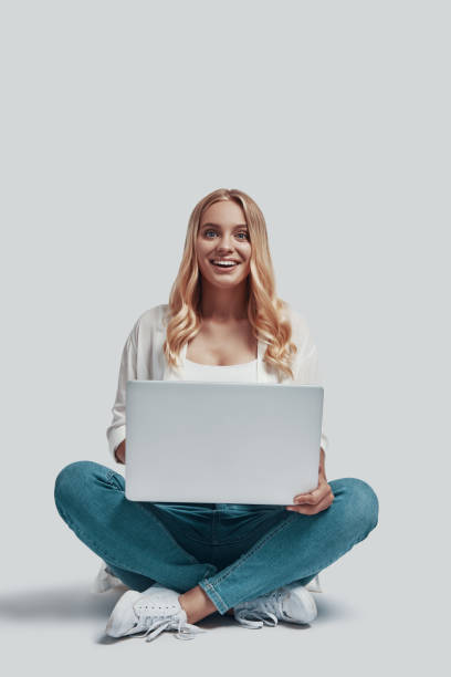 istock Attractive young woman using laptop and smiling while sitting against grey background 1181204447
