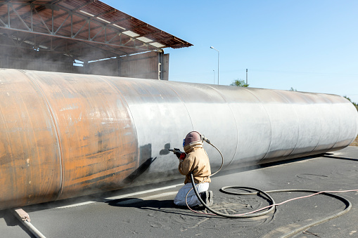 View of the manuel sandblasting to the large pipe. Abrasive blasting more commonly known as sandblasting is the operation of forcibly propelling a stream of abrasive material against a surface under high pressure.