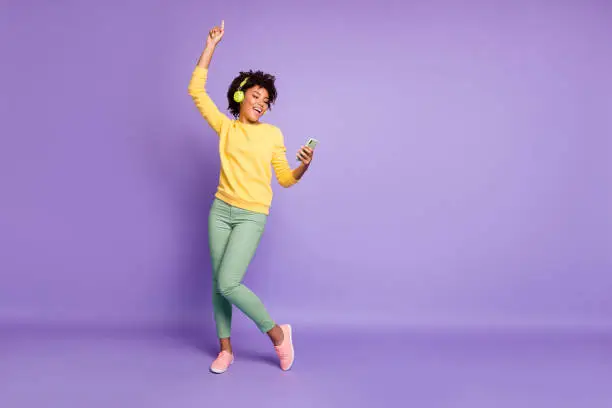 Full length body size photo of curly wavy trendy sweet cute charming, carefree girl dancing and singing while reading song text on her telephone and wearing headphones isolated violet pastel color background