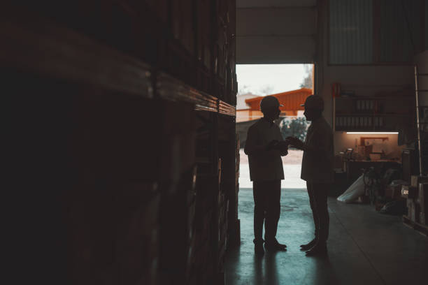 istock Silhouette of two hardworking warehouse workers standing in warehouse and checking inventory. Older one holding tablet while younger one looking at it. 1181214970