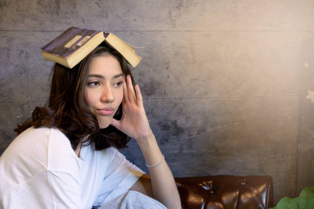 Young woman bored face with book on head sitting on sofa in the room with morning light background.