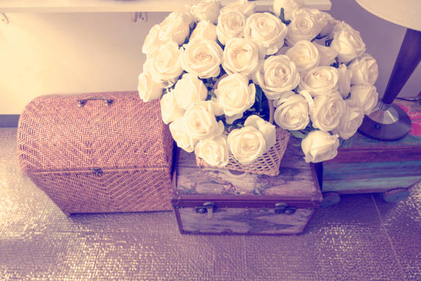 White roses in basket on wooden box and bamboo bag decoration on the floor indoor with wall background.
