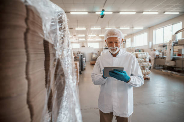 istock Smiling Caucasian senior adult inspector dressed in white uniform using tablet for quality assessment of food in food plant. 1181216015