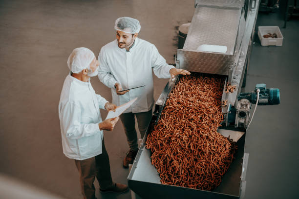 Top view of two Caucasian quality controllers standing next to machine with salty sticks and evaluating quality. Both are dressed in white uniforms and having hairnets. Food plant interior. vista superior de dos controladores de calidad caucásicos de pie junto a la máquina con palos salados y la evaluación de la calidad. ambos están vestidos con uniformes blancos y con redes para el cabello. interior de la planta de alimentos.