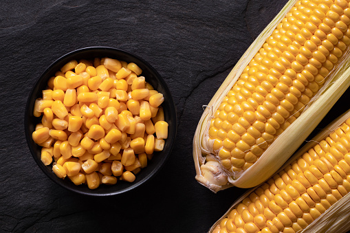 Canned sweet corn in a black ceramic bowl next to two corn cobs in husks on black slate. Top view.