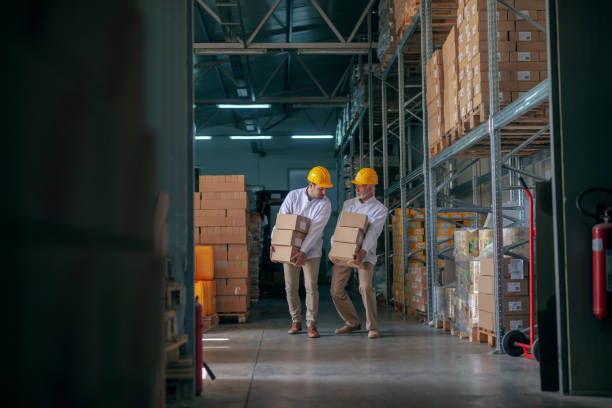 istock Two storage workers dressed in white uniform and with yellow helmets carrying heavy boxes. 1181217972