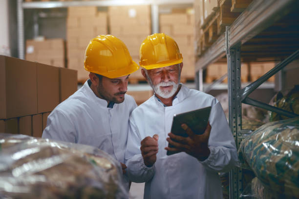 istock Two hardworking warehouse workers standing in warehouse and checking inventory. Older one holding tablet while younger one looking at it. 1181218332