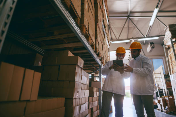 istock Two hardworking warehouse workers standing in warehouse and checking inventory. Older one holding tablet while younger one looking at it. 1181218382