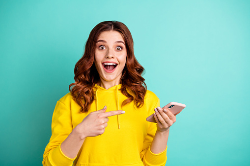 Photo of beautiful cheerful ecstatic attractive curly wavy trendy stylish, youngster pointing at telephone held with hands wearing yellow sweater hoodie isolated over vivid teal color background